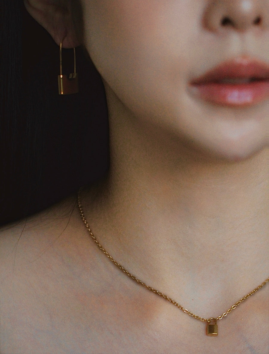 Close-up of a person wearing mini gold earrings and a mini lock pendant necklace with a dark background